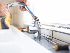 Hands removing a chrome faucet spout from a kitchen sink for repair or replacement.
