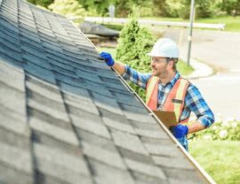 Inspector on roof edge wearing hard hat and vest checking shingles with clipboard