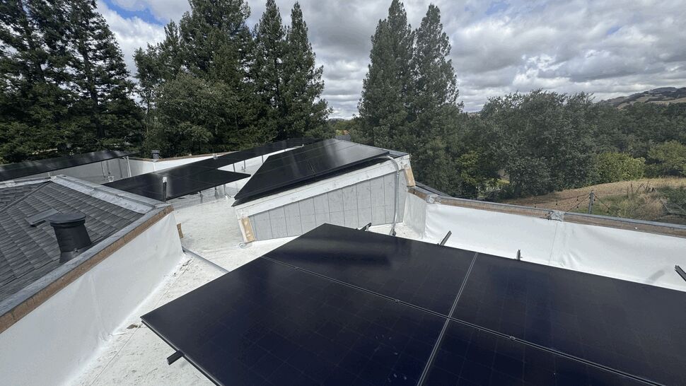 Multiple solar arrays on a flat section of roof with tall trees and hills beyond.