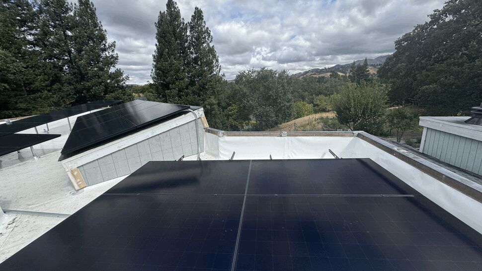 Panoramic Solar Installation | Green Air Heating & Air Conditioning Inc Solar panels on a rooftop overlooking a valley and tree-covered hills under a cloudy sky.