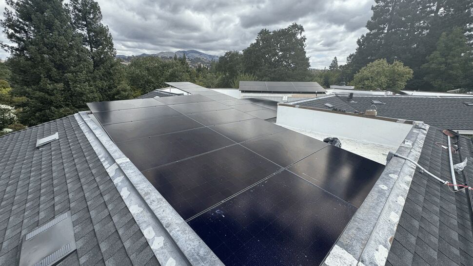 Black solar panels installed on a sloped rooftop with trees and cloudy sky.