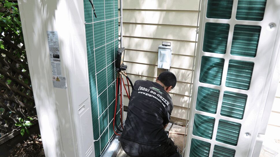 Technician kneeling between two outdoor condenser units with gauges and hoses