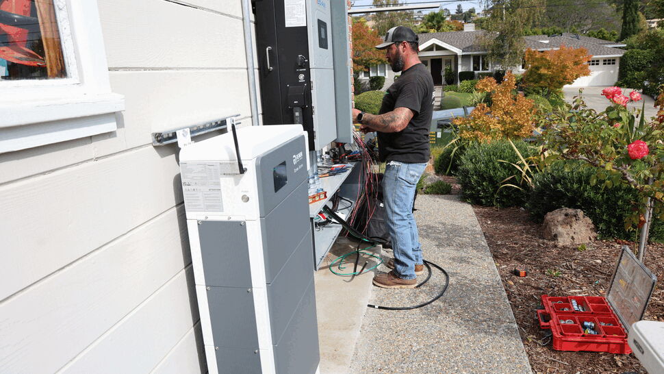 Solar Storage Wiring | Green Air Heating & Air Conditioning Inc Side view of technician connecting wires to solar storage components near landscaping