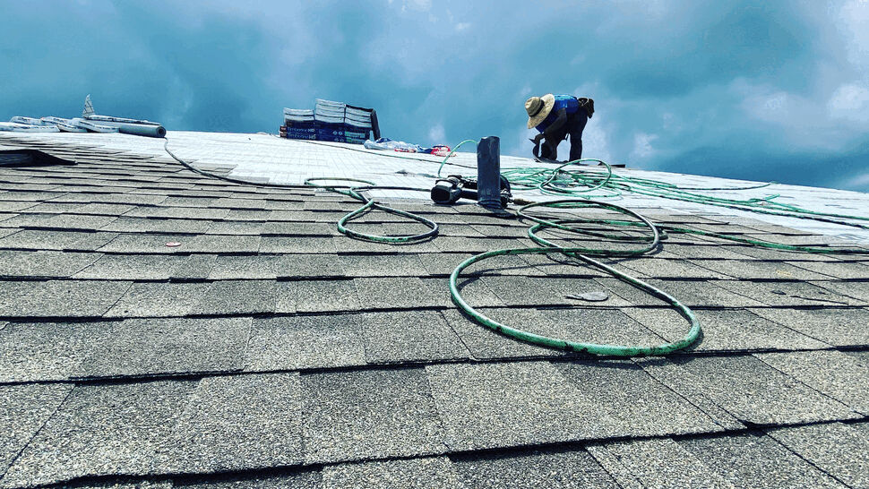 Roofer kneeling on shingled roof with tools, hoses, and cloudy sky overhead