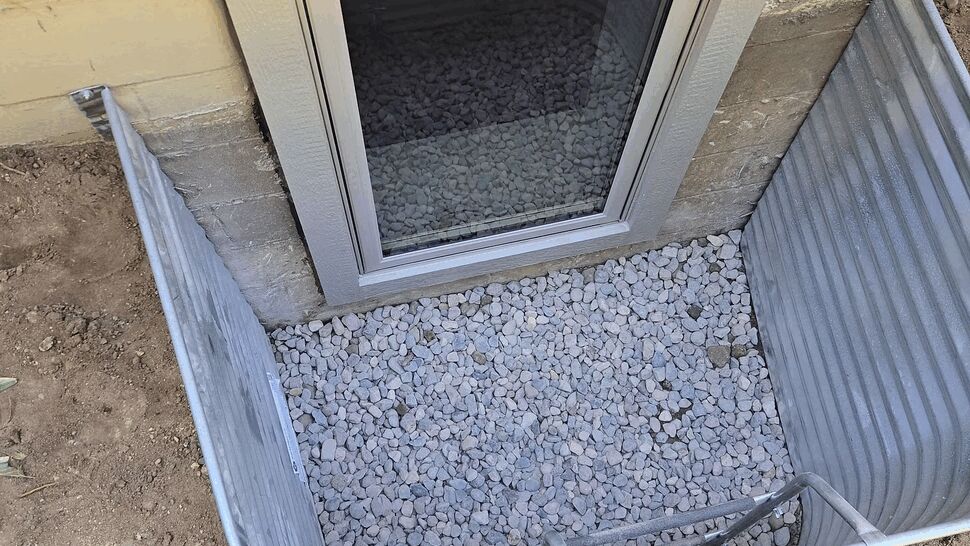 Top-down view of a basement window well filled with gravel and a framed window.