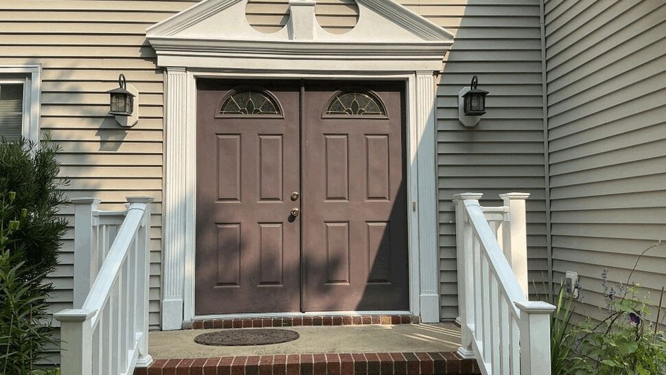 Brown double front doors with brick steps, white railings, and beige siding