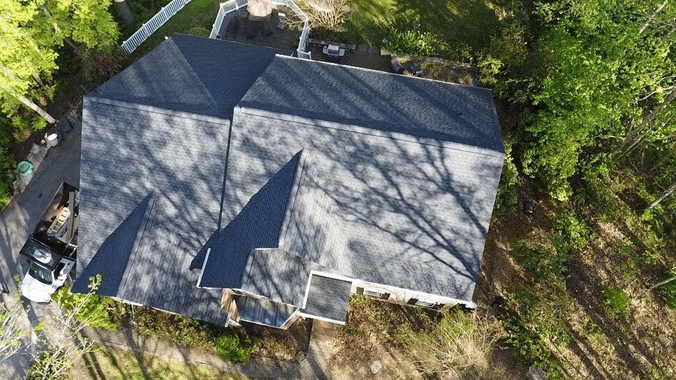 Top-down aerial of a dark shingled roof with tree shadows and work trucks at the driveway.