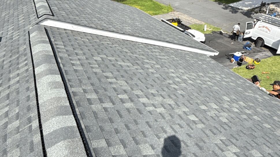 Close-up view of gray shingle roof with ridge vent and workers below