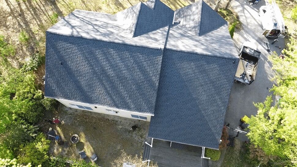 Drone view of a dark residential roof with on-site equipment and trucks nearby.
