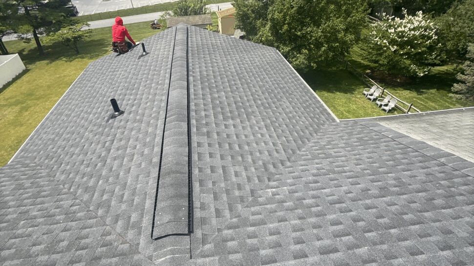 Wide aerial shot of a full gray shingle roof with crew member and surrounding trees