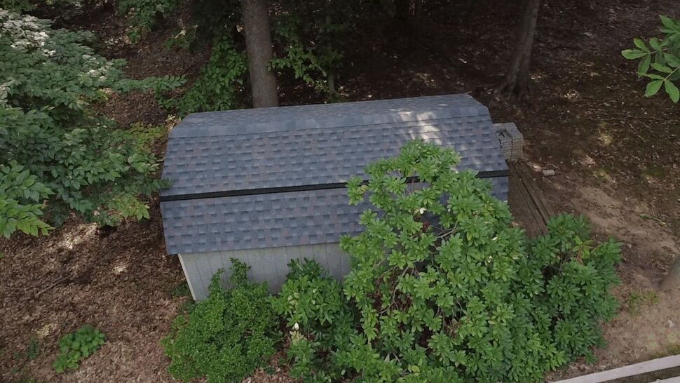 Aerial Storage Shed Roof Inspection | Wells Home Improvements Aerial view of a small shingled storage shed roof surrounded by trees.